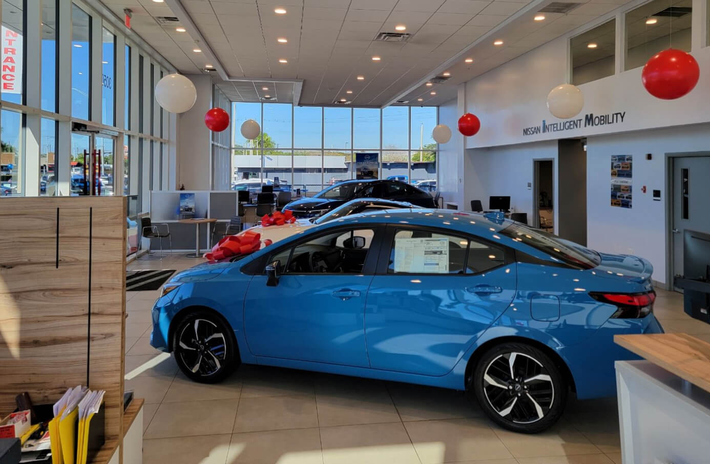 Profile view of a blue Nissan Versa on a dealership showroom floor with decorative balloons.