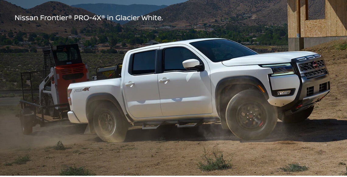 Dynamic front three-quarter tracking view of a Nissan Frontier PRO-4X in Glacier White pulling a loaded utility trailer.