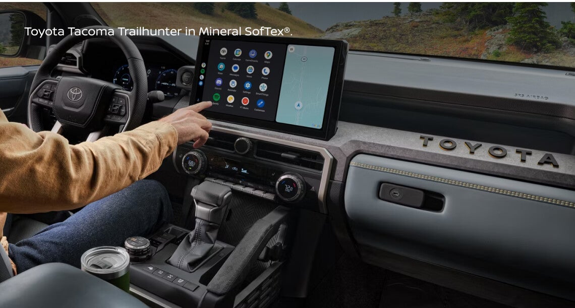 Interior dashboard view of a 2025 Toyota Tacoma Trailhunter in Mineral SofTex® showing the driver operating the central infotainment touchscreen.