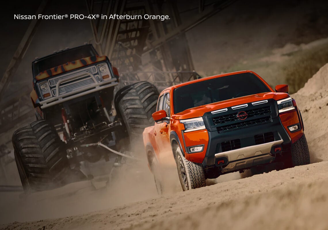 Dynamic front-angle tracking view of a Nissan Frontier PRO-4X in Afterburn Orange towing a custom monster truck.